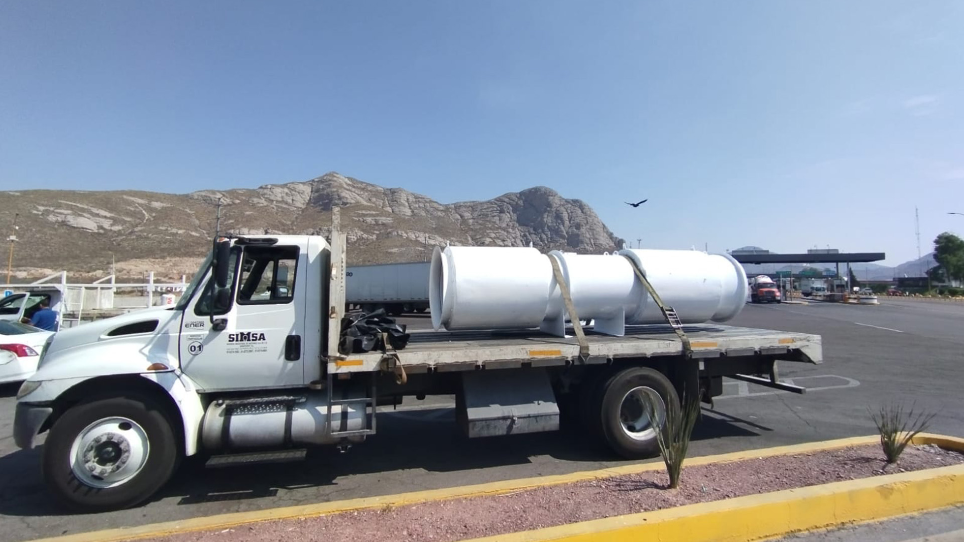 Camión de plataforma blanca de la empresa SIMSA transportando un ventilador industrial axial de gran tamaño en una carretera de zona montañosa.
